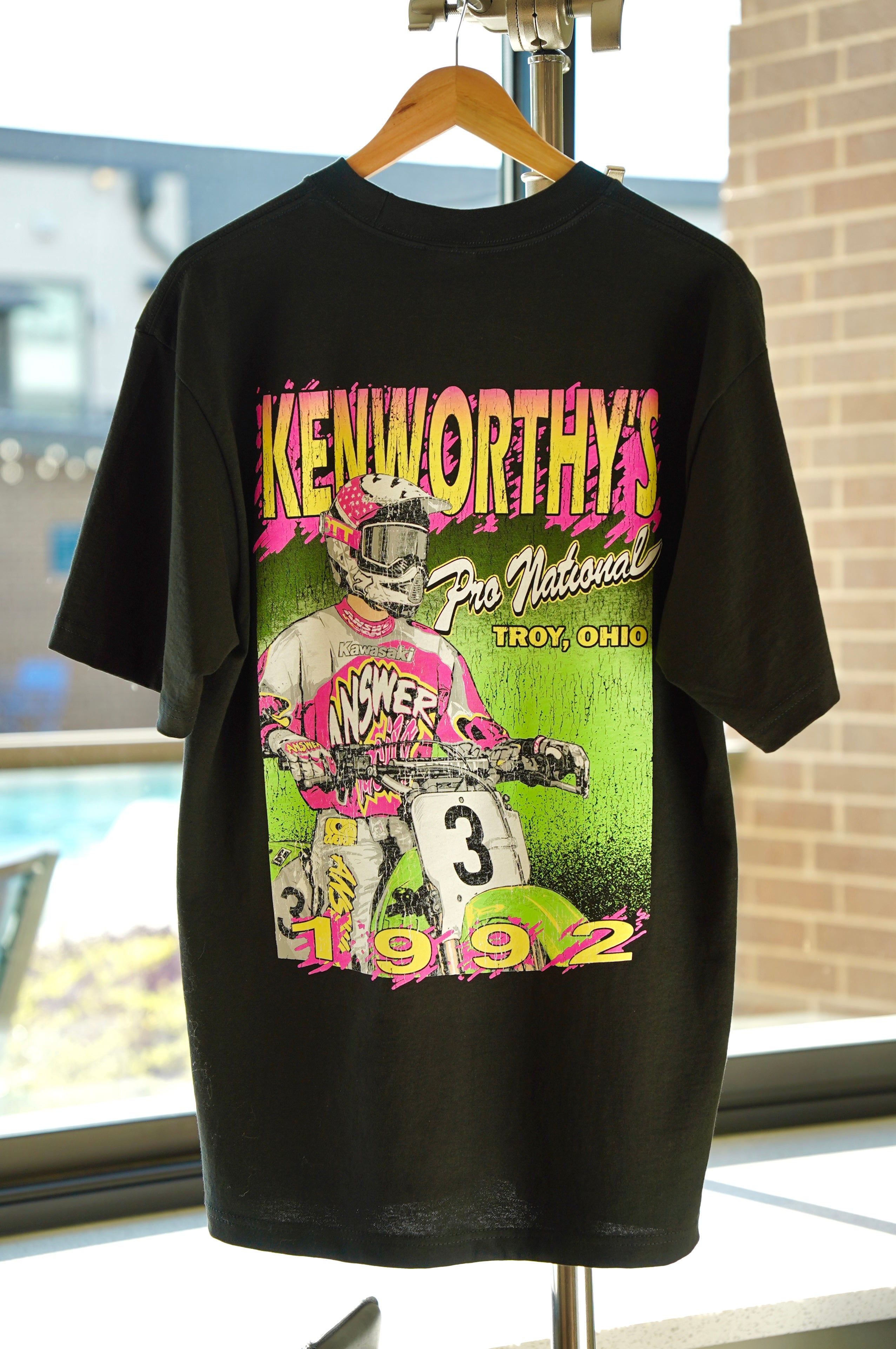 A black short-sleeve graphic t-shirt hanging on a wooden hanger. The shirt features a large, colorful front print with the text 'KENWORTHY'S Pro National Troy, Ohio 1992' and an illustration of a motocross racer in pink and white gear against a green background.